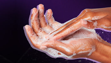 A person rubbing hands with foamy cleanser under a running faucet in a white tiled bathroom.