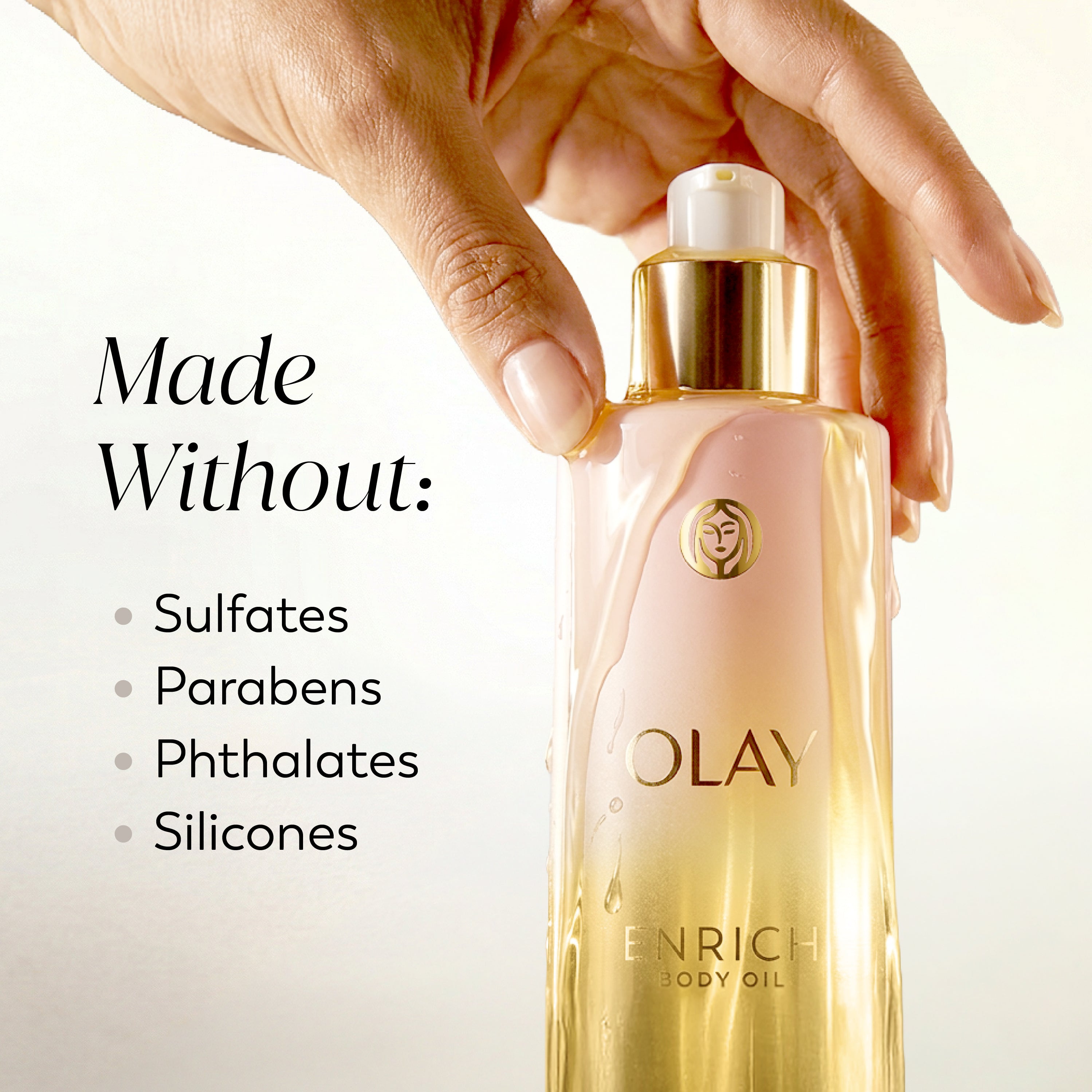A hand holds a bottle of Olay Enrich Body Oil against a clean white background, with text on the left reading 'Made Without: Sulfates, Parabens, Phthalates, Silicones.'.