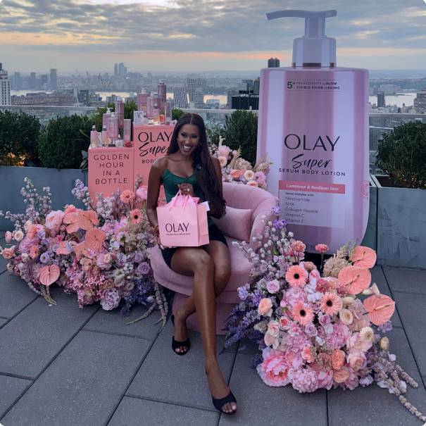 Person seated at a pink Olay display, holding an Olay bag and surrounded by flowers and Olay products.